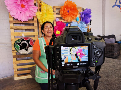A Latina woman standing in front of a camera. The camera viewfinder is shown in the foreground.