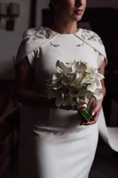 closeup of a bride wearing a dress with embroidered capped sleeves holding a bouquet of calla lilies in a bedroom at the nomad hotel before her bespoke wedding