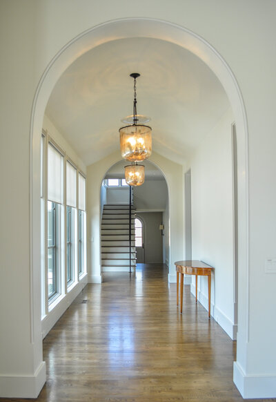 Image of white hallway leading to staircase with arched doorways, clean lines, and beautiful chandeliers