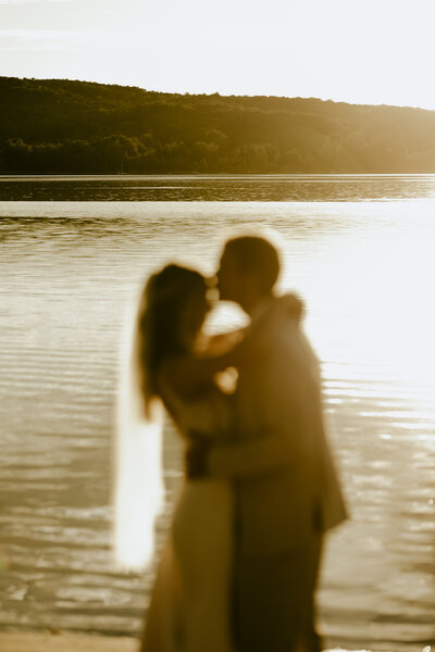 A groom kissing his wife's forehead in front of a lake at sunset