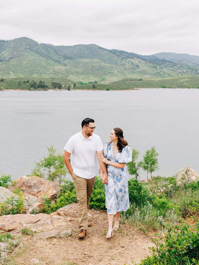A couple walks hand in hand on a rocky path by a lake, surrounded by greenery and mountains under a cloudy sky—captured beautifully by a Colorado Wedding Photographer.