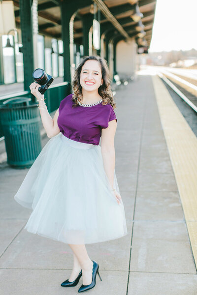 woman in a purple and white dress holding a camera with one hand at a train station