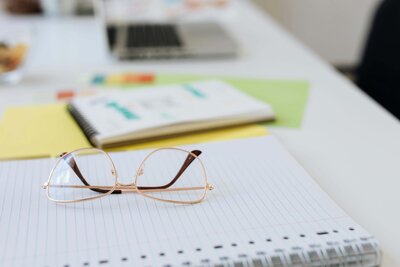 A pair of gold-framed glasses rests on a lined notebook, surrounded by other stationery items and a blurred laptop in the background on a tidy workspace.