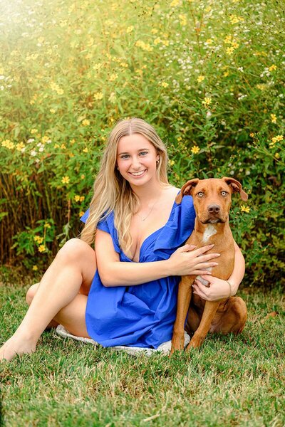 charlotte grad wearing a blue dress sits beside her brown dog in a flower patch