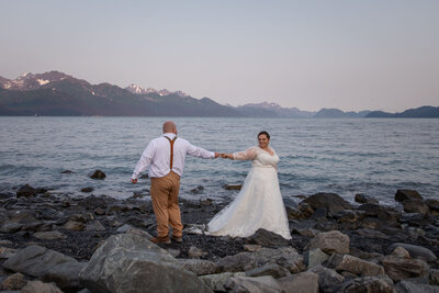 A bride in a white dress walks away from her groom on a beach in Alaska and reaches back to touch his hand.