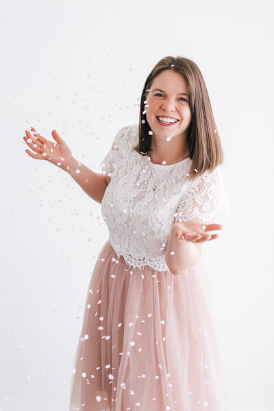 colorado wedding photographers headshot with photographer in a blush pink tulle skirt and lace top throwing confetti in the air and smiling on a white backdrop