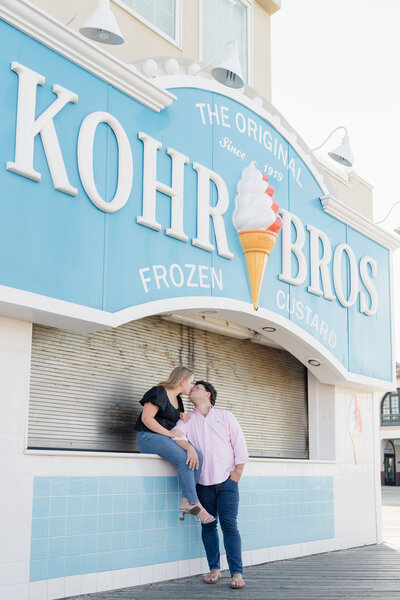Engaged couple kisses in front of Kohr Bros during sunrise engagement session on the boardwalk in Ocean City, NJ