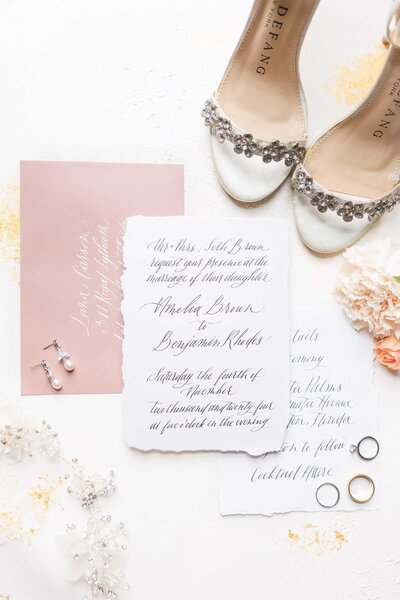 hand written invitation suite, pink calligraphy envelope, flowers, wedding shoes, and wedding rings  in a flat lay image