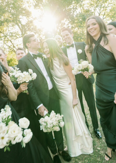 Off center photo of bride and groom surrounded by smiling wedding party with sun shining through
