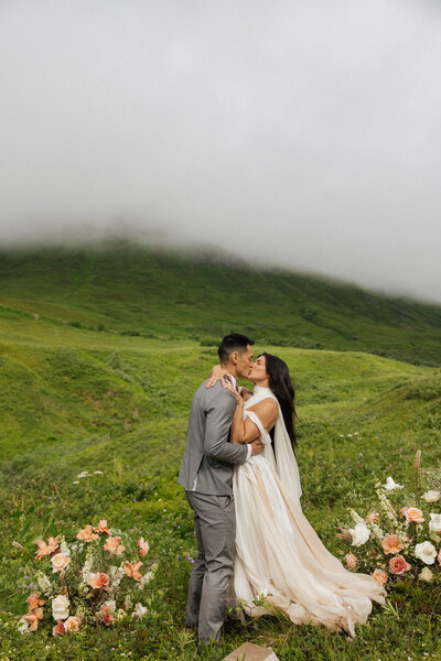 bride and groom kissing standing on a rocky shoreline with tall black rocks in the background and trees