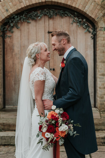 Bride and groom smiling holding autumnal orange and red bridal bouquet at their wedding at The Ravenswood wedding venue