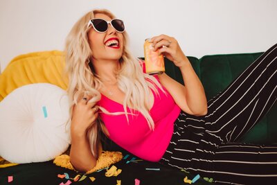Andi Sweeny laying on a green couch wearing black and white pin stripe pants and a bright pink shirt and sunglasses throwing her head back in laughter.