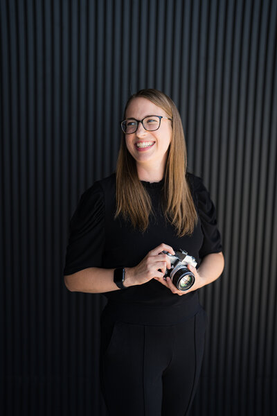 Grace from Sam & Grace standing smiling holding a camera in front of a black neutral wall