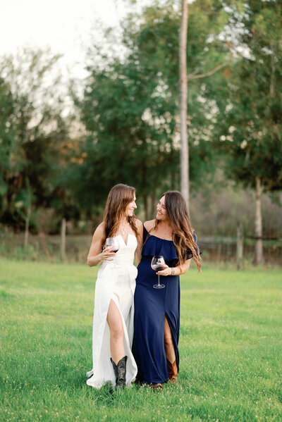 Two LGBTQ wives in classy dresses holding wine glasses walking together through the grass while looking at each other and smiling
