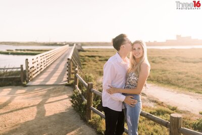 Bride to be receives a kiss from her Groom on the side of her head as they embrace each other during photo shoot at the Bolsa Chica Ecological Reserve in Huntington Beach