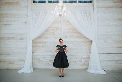 A wedding officiant wearing a black dress holding a leather bound book standing beneath a ceremony arbor made of sheer white fabric in an all white wedding barn