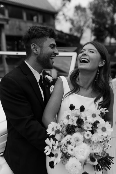 Newlyweds sharing heartfelt laughter in a wedding photoshoot by a vintage car, creating timeless memories.