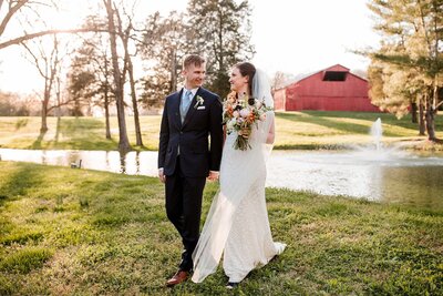 Bride and Groom walking by pond with red barn in the background at Cedarmont Farms