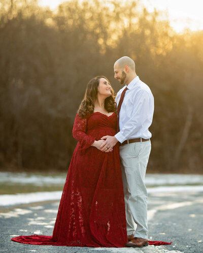 A pregnant woman in a red lace maternity gown and a man wearing a dress shirt and slacks stand in a snowy road at sunset, looking at each other,