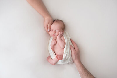 Baby sleeping in white wrap with parents hands around him at his Detroit Newborn Photography session