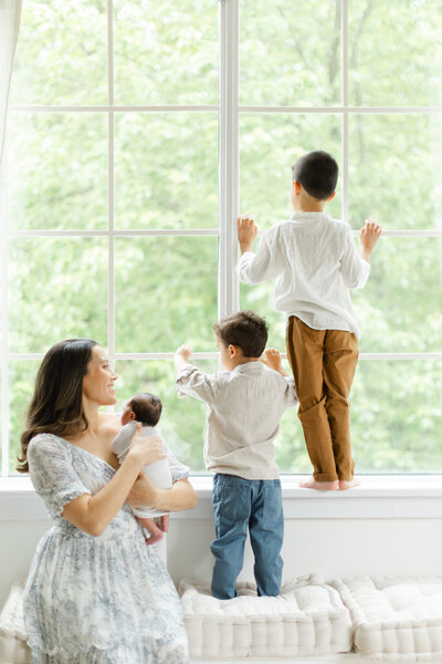 Mother holds newborn baby as older sons look out the photography studio window