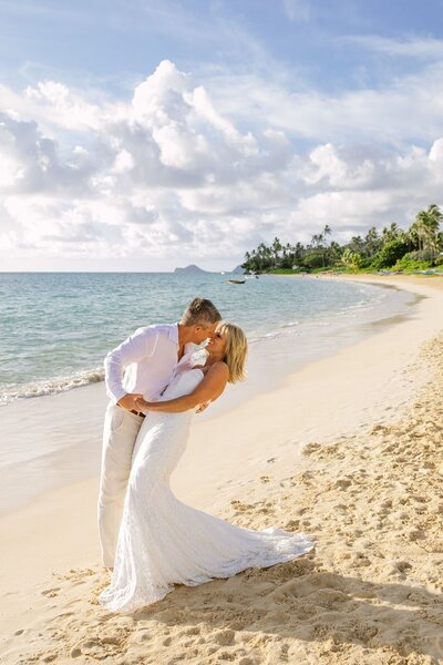 A man holds and dips a woman wearing a wedding dress on the beach.