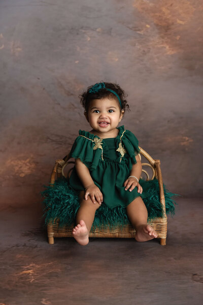 A happy toddler girl in a green dress sits on a wicker bench in a New Orleans Family, Maternity & Newborn Photographer studio