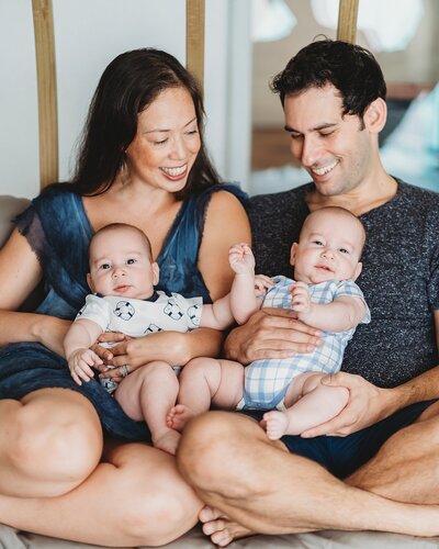 A joyful family sitting together; a woman and a man hold two smiling babies on their laps, all looking delighted and content.