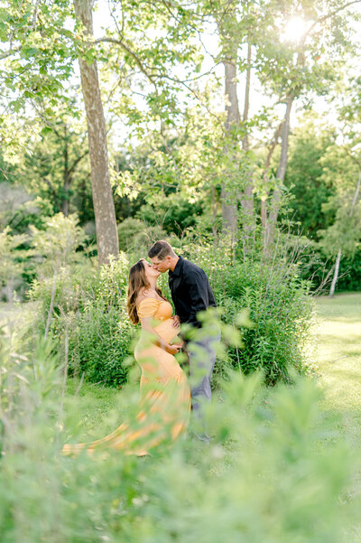 pregnant woman kissing her husband in a beautiful lush garden with sun streaming in.