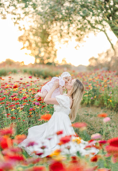 A mommy and me photography session by Bay area photographer shows a mom holding her baby in a white jumper sitting in a field of flowers.