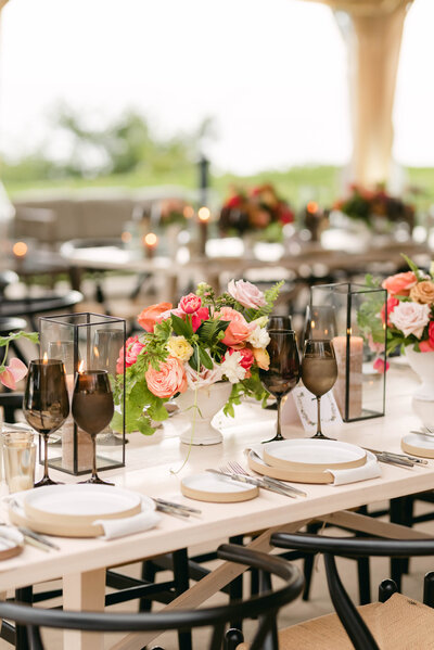 Wedding reception: a cream coloured table, wooden black chairs, black wine classes, cream dishes, black lanerns, and red flowers
