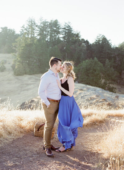 a couple on a mountain top, wind in their hair. she is in a blue skirt and he is in tan pants. they are on mt tam in San Francisco.
