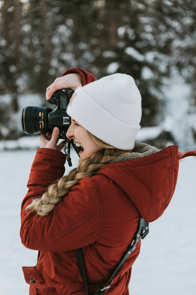 a photographer smiling and taking a photo wearing a red carhartt jacket with blonde hair in a braid and a beanie