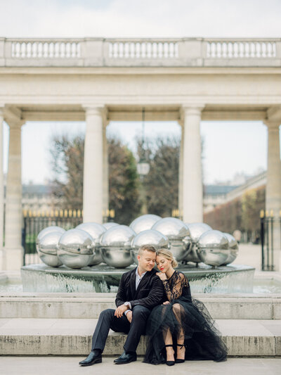 a man and woman in a black suit and black tulle dress sitting on some steps in front of giant silver ball statue at palais royal in paris