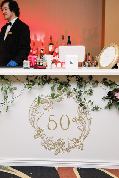 Custom acrylic monogram in gold attached to the front of a bar at a 50th anniversary celebration. The stylish monogram adds a sophisticated touch to the bar area, complementing the overall theme of the event.
