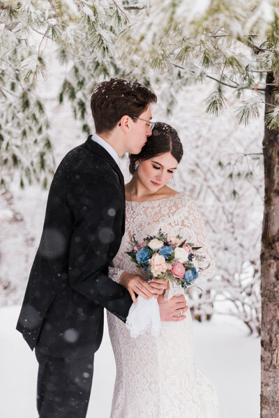 winter Rocky Mountain National Park wedding with bride and groom embracing in a snowy woodland area while the bride holds her wedding bouquet