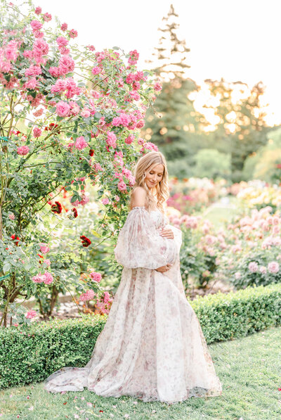 A maternity session photographed by Bay area photographer shows a woman dressed in a light and airy gown holding her baby bump standing in a rose garden.