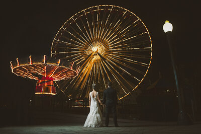 Bride and groom stand in front of a ferris wheel at an amusement park