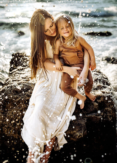 Family photographer, a mother and daughter sit on rock at the beach with crashing waves behind them