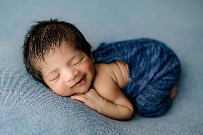 dark hair baby boy is smiling as he is posed laying on his stomach in a blue outfit on a blue background