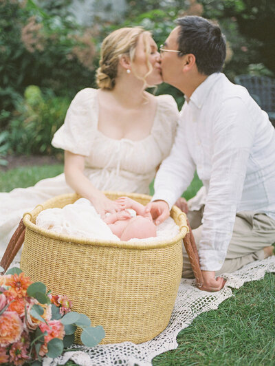 A Maryland newborn session where a mother and father sit outside in a garden and lean in for a kiss while placing a hand on their baby in a Moses basket.