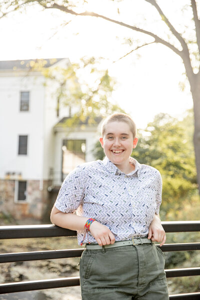 high school senior stands on bridge with bright light and mill behind, smiles