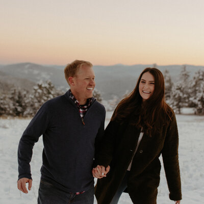 The couple is running through the snow laughing with each other as the sun sets under the mountains.