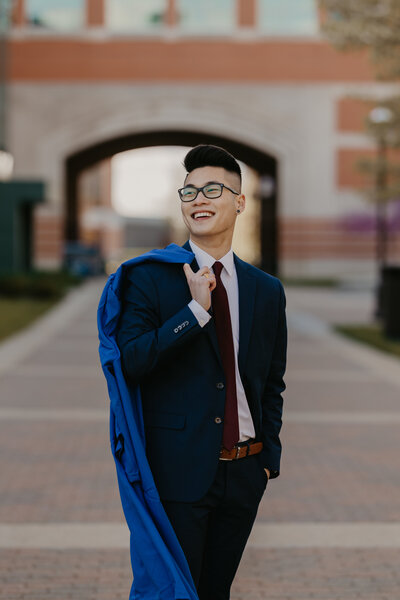 Man posing in front of GVSU campus buildings with his graduation gown over his shoulder, looking into the distance and laughing.