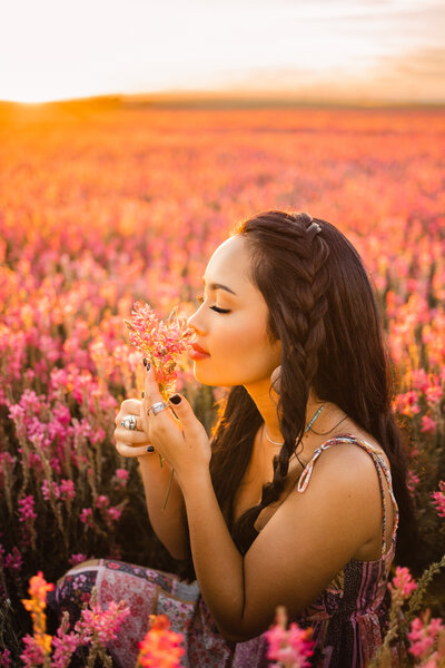 senior girl with dark hair sitting in a field of pink flowers at sunset