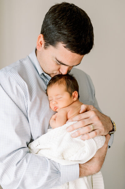 Dad in blue shirt holds baby boy with dark hair in white blanket and kisses the top of his head