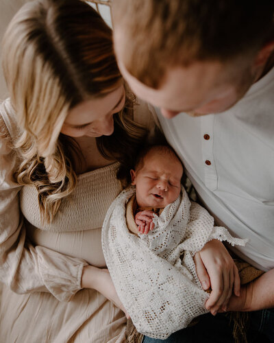 Sweet newborn photography photoshoot captured by Morgan Ashley Lynn Photography in Milwaukee, WI, closeup and overhead view of a mom and dad holding their newborn boy on their laps, wrapped up in a vintage white floral blanket, smiling