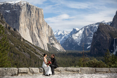 A bride wearing a white dress and holding a bouquet of red flowers sits on a rock wall in Yosemite facing her groom  who wears a black suit.