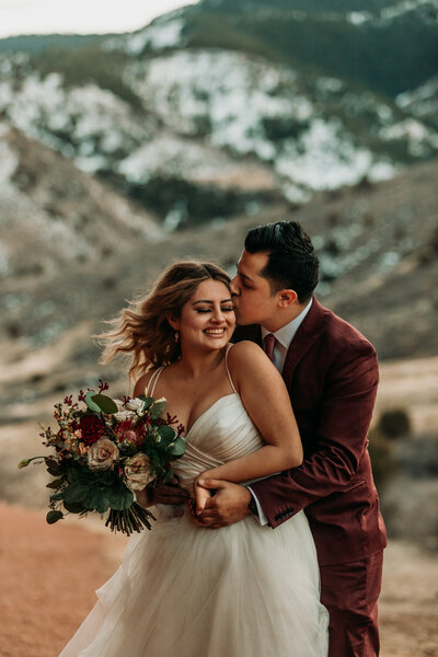 Groom tenderly holds  and kisses smiling bride with bouqet in front of Red Rock Mountain Range