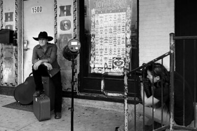 Country music photography behind the scenes Mark Maryanovich sitting on stairs while photographing Darrell Goldman sitting on suitcase guitar leaning beside him black and white image